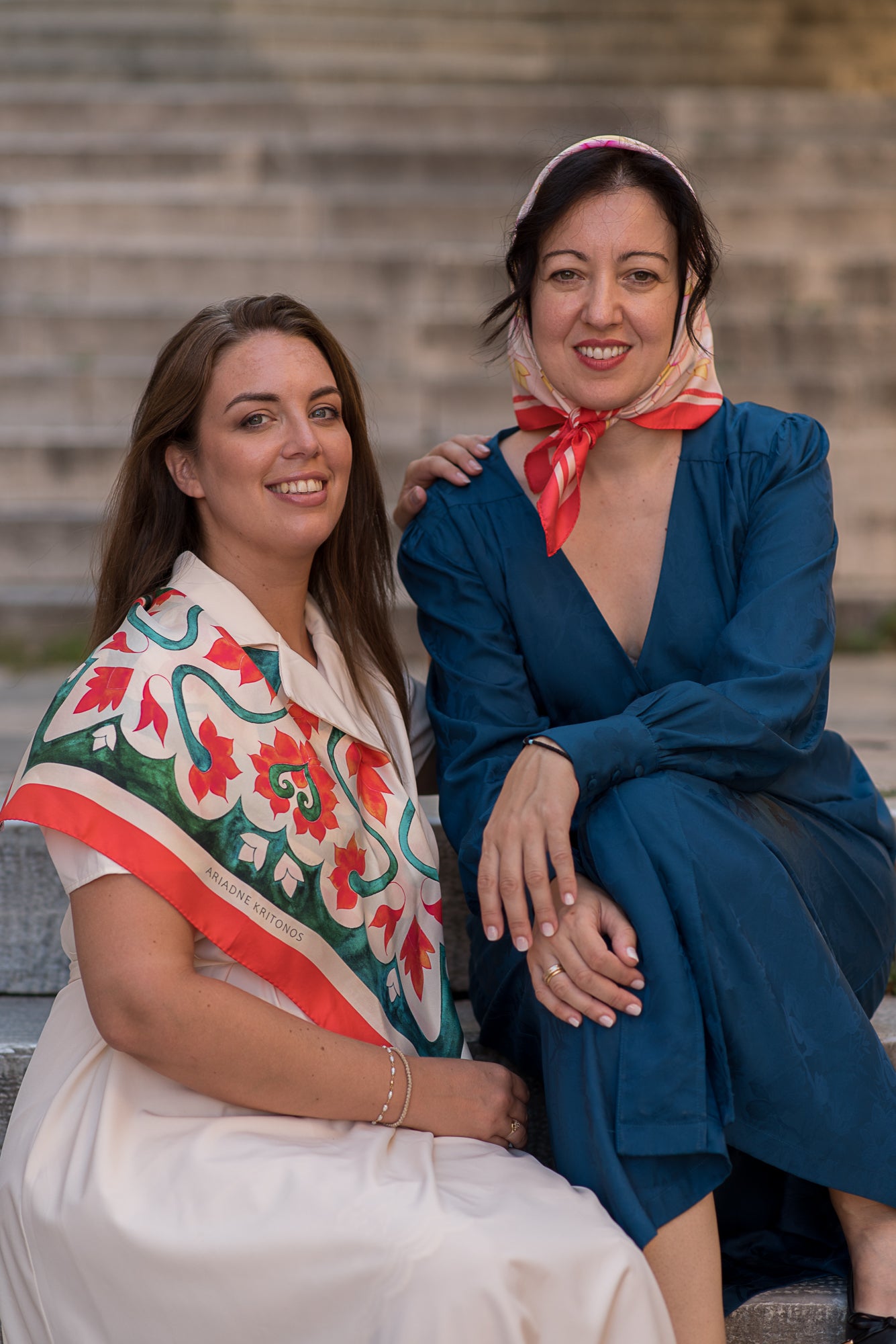 Two women sitting on steps wearing the Teal and Pink Attica Garden silk scarves and smiling