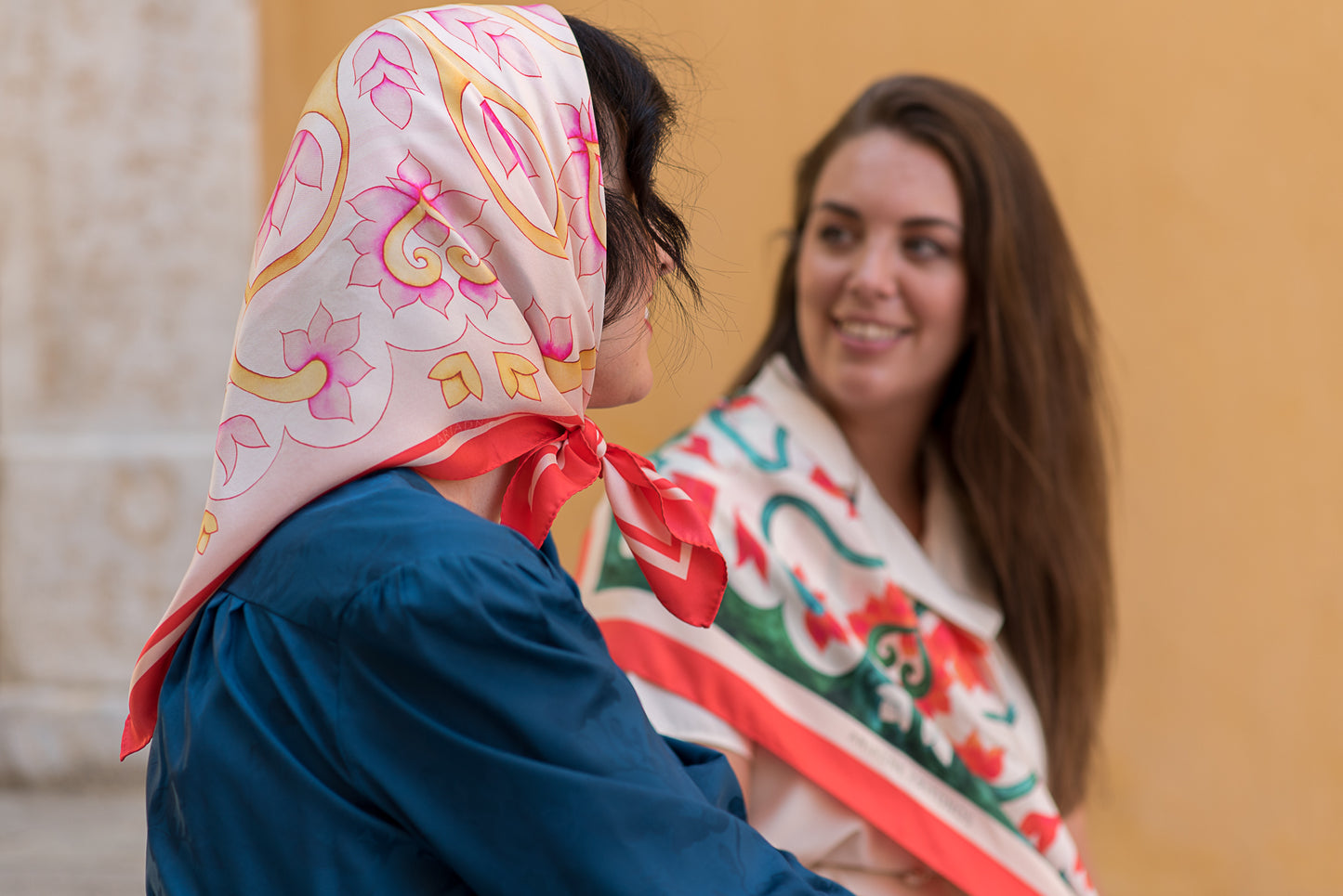 Two women wearing the pink and teal Attica Garden silk scarves styled on the head and shoulder