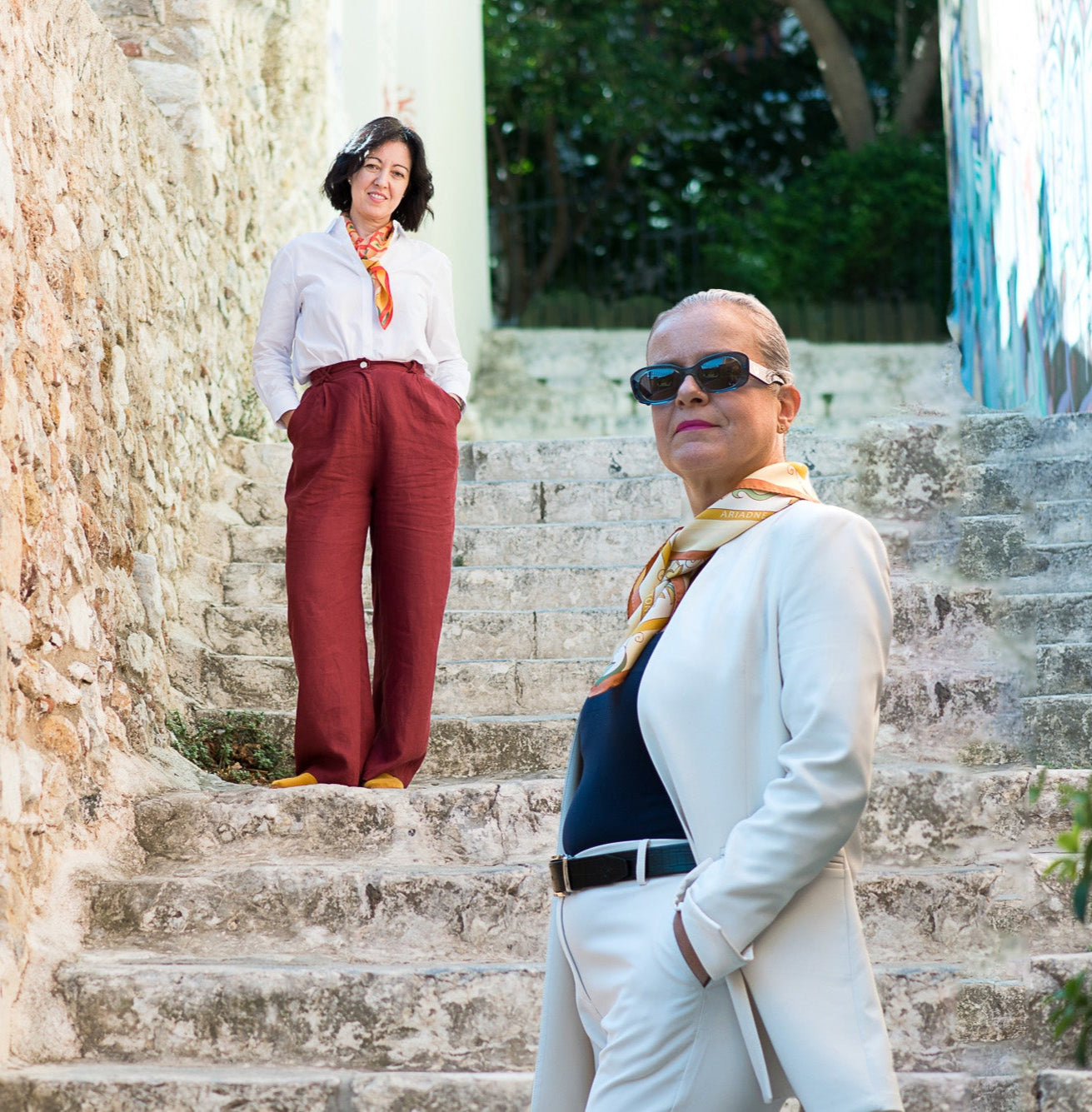 Red and cream Mesogeia Blooms silk scarves worn by two women standing on stone steps next to a stone wall
