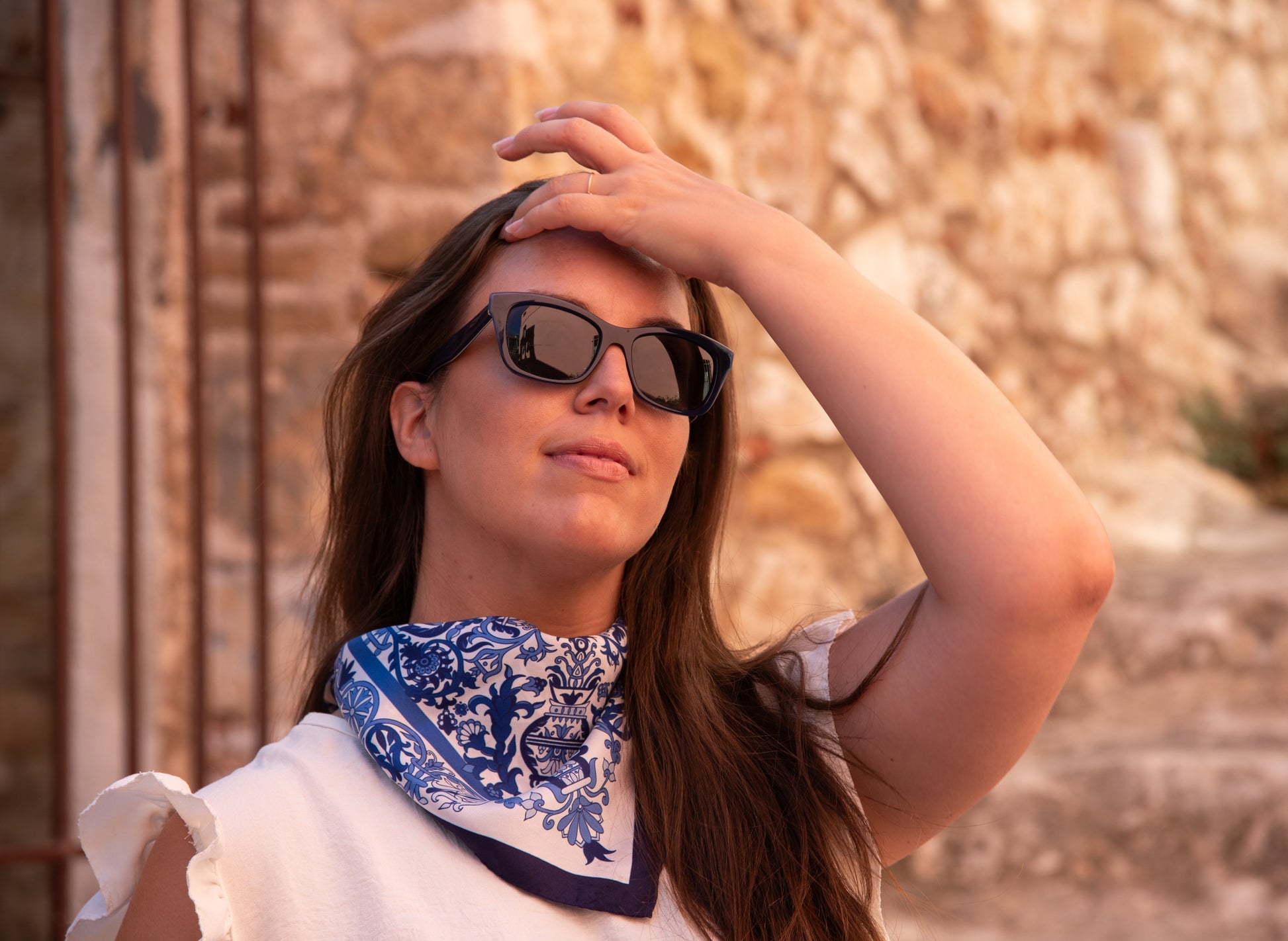 Model wearing sunglasses is showing her Cretan Mermaid silk scarf in blue styled as neckerchief