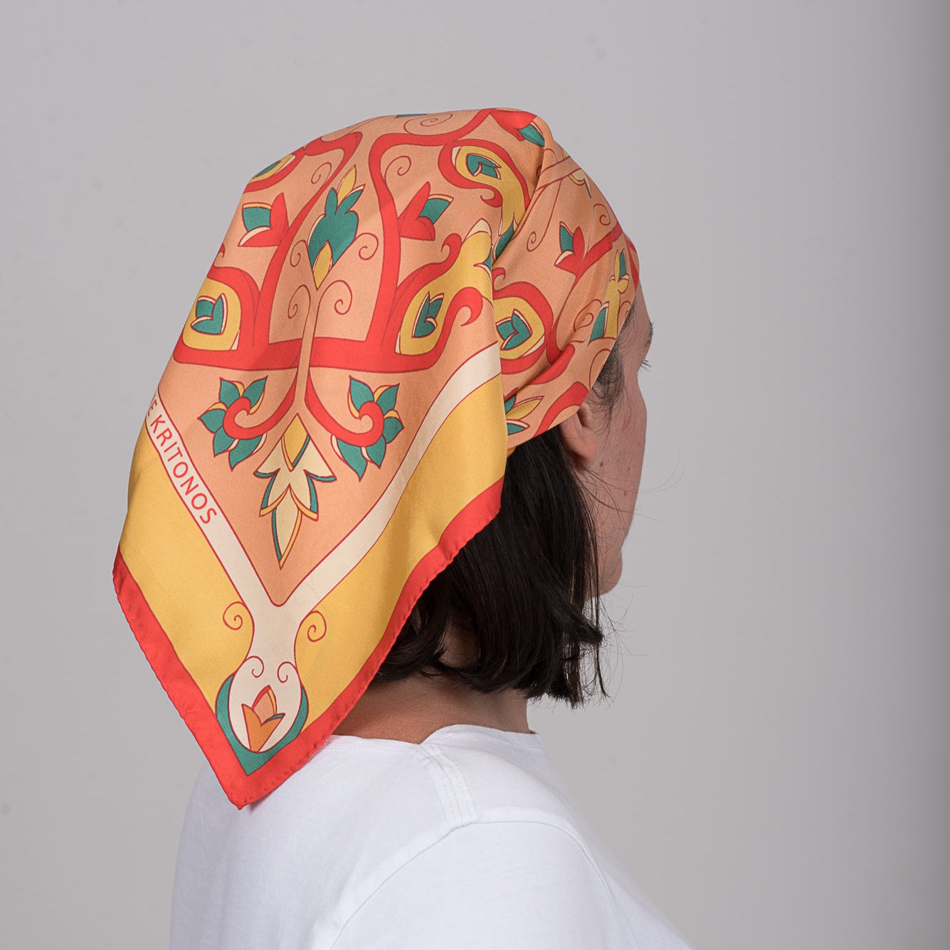 The red Mesogeia Blooms square silk scarf in warm orange tones worn as head wrap