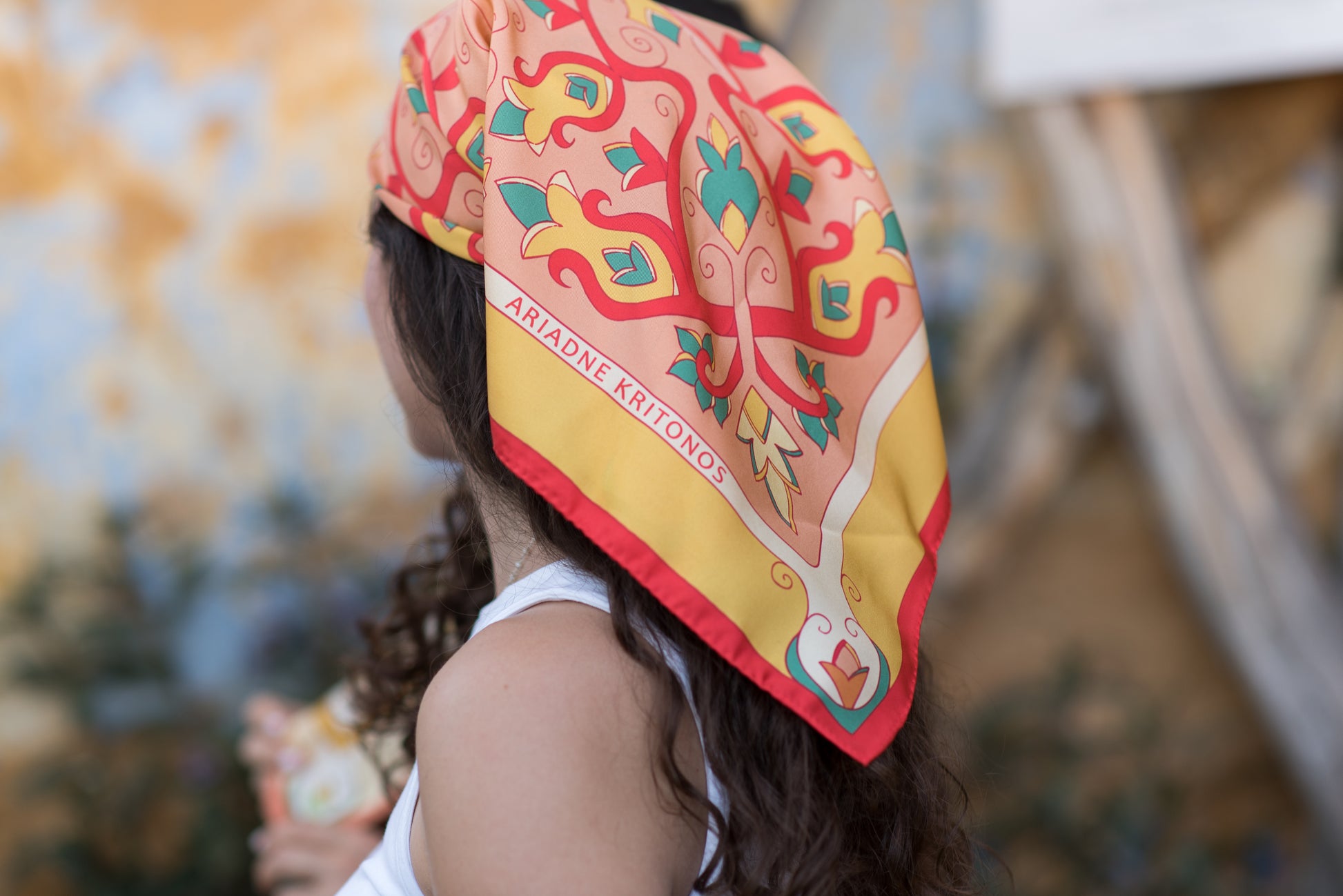 Model wearing red and pink Mesogeia Blooms silk scarf as headscarf and showcases the brand logo which reads Ariadne Kritonos