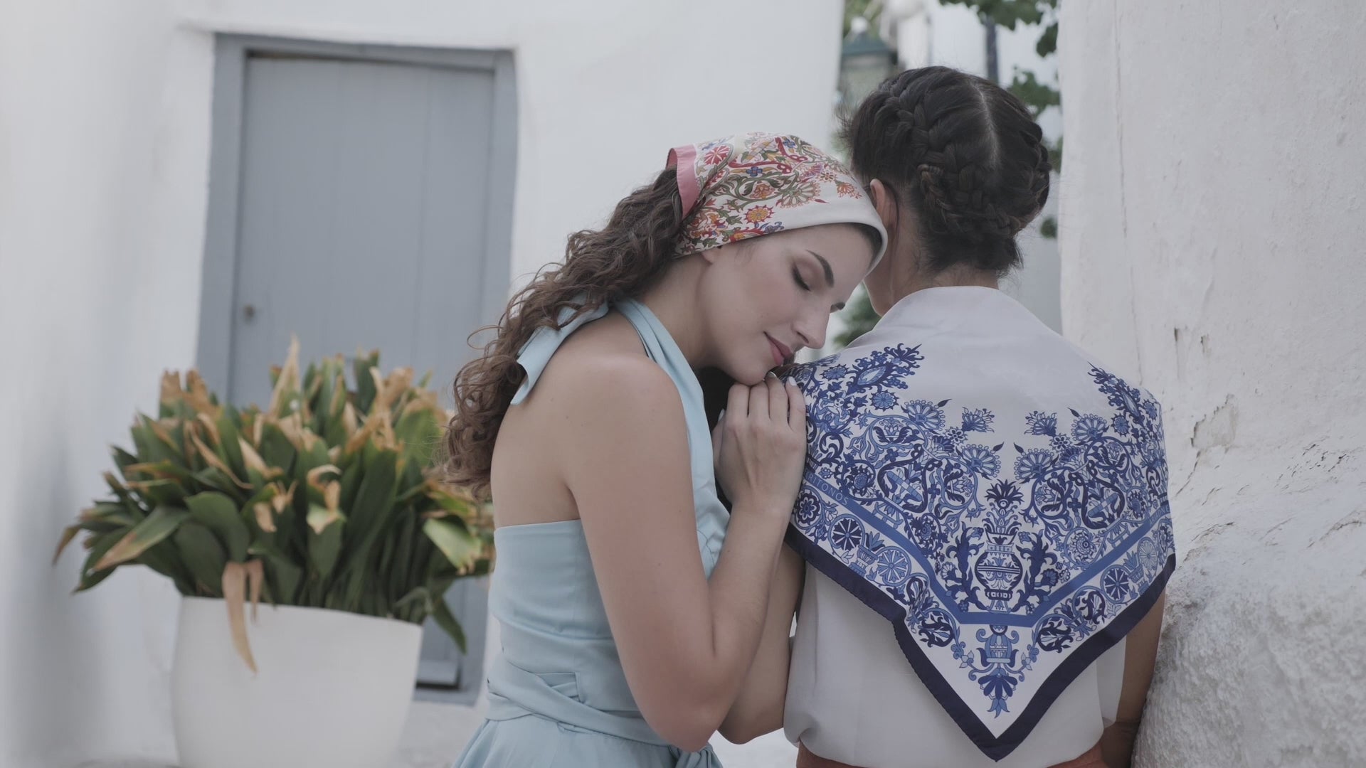 Close up video of model wearing blue Cretan mermaid silk scarf on her back
while another model rests her head on her shoulder and wears the pink version of the scarf.
